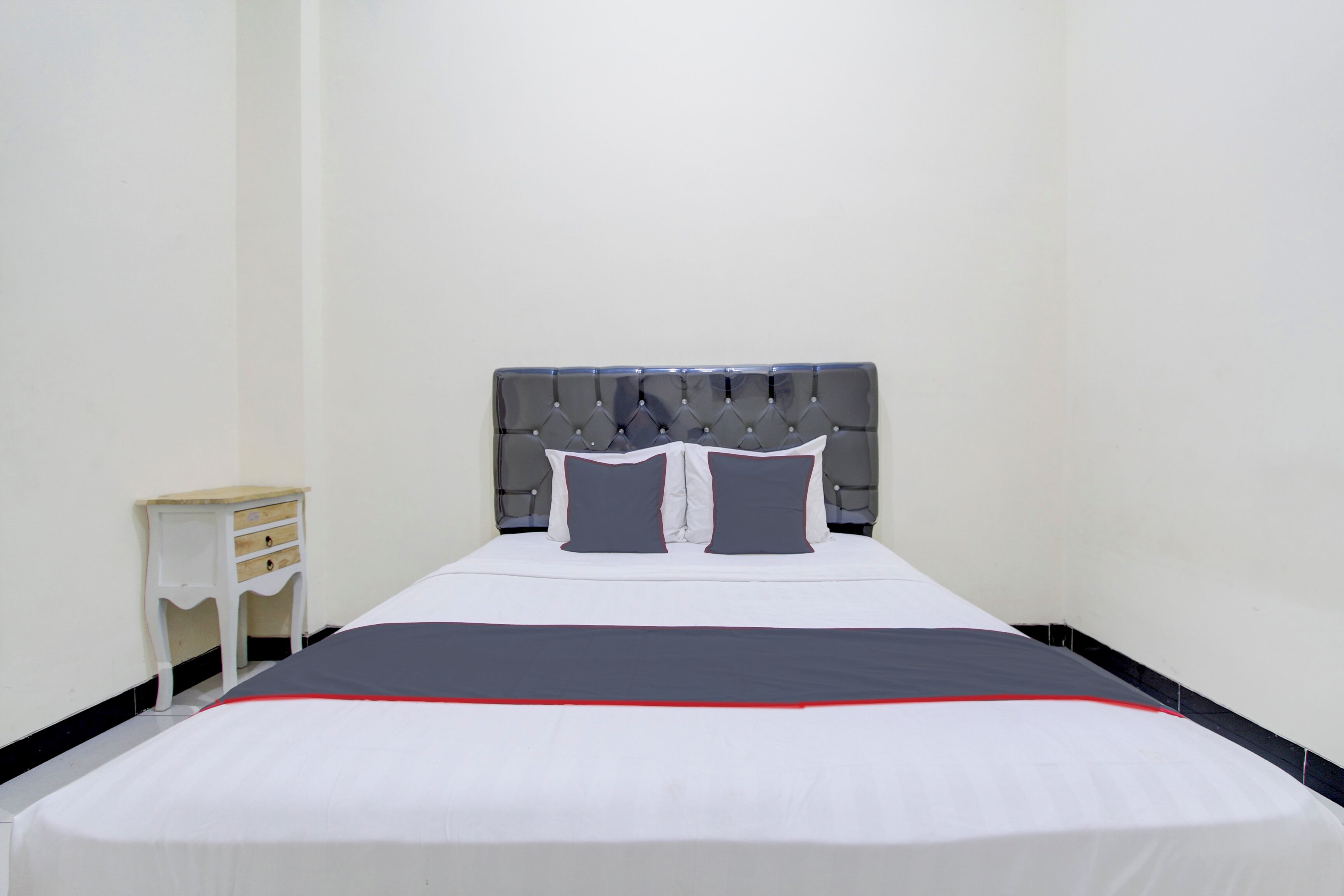 Una cama bien hecha con un cabecero tapizado en gris, ropa de cama blanca y un detalle rojo en la colcha.