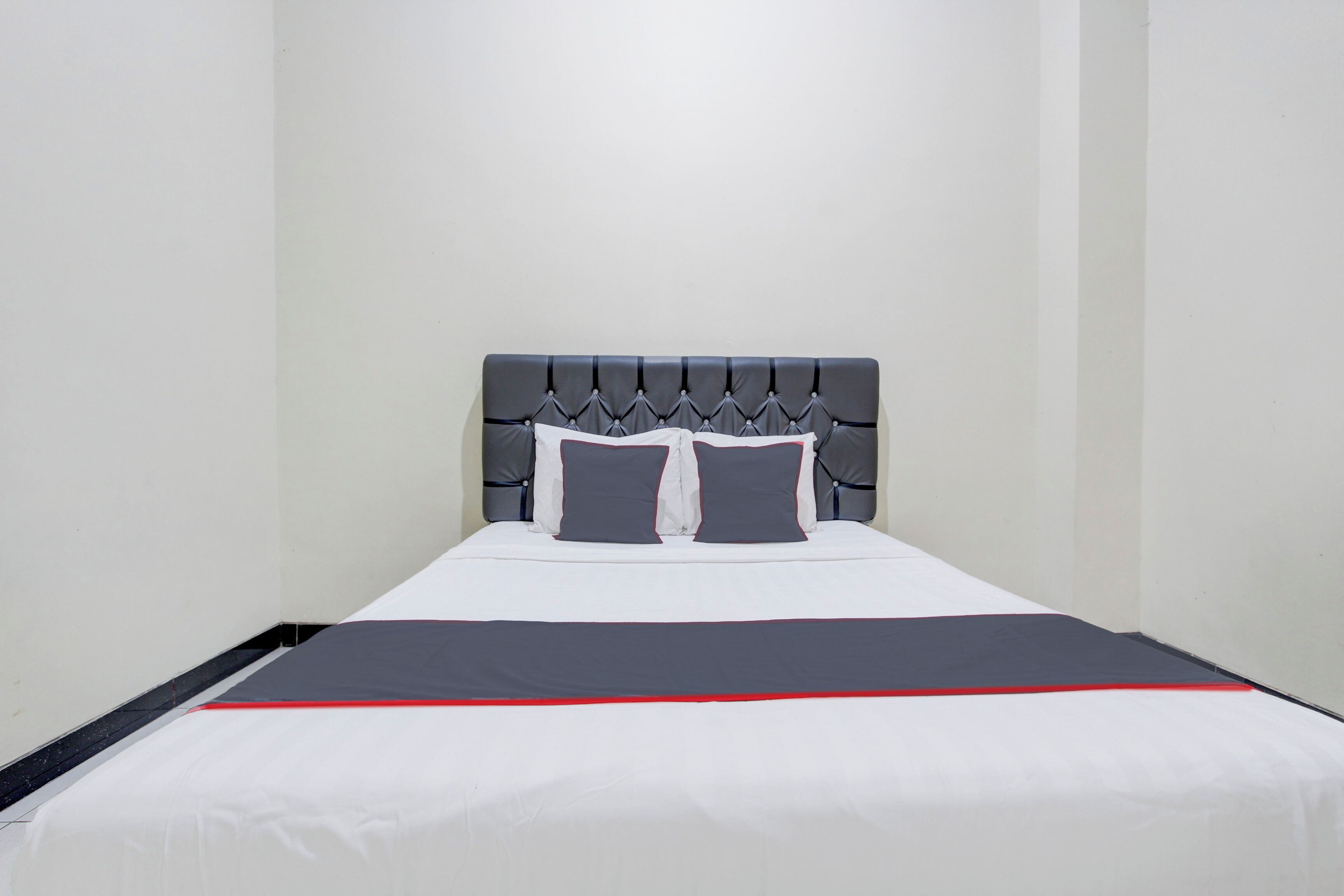 A neatly made bed with a grey upholstered headboard, white bedding, and a red accent strip.