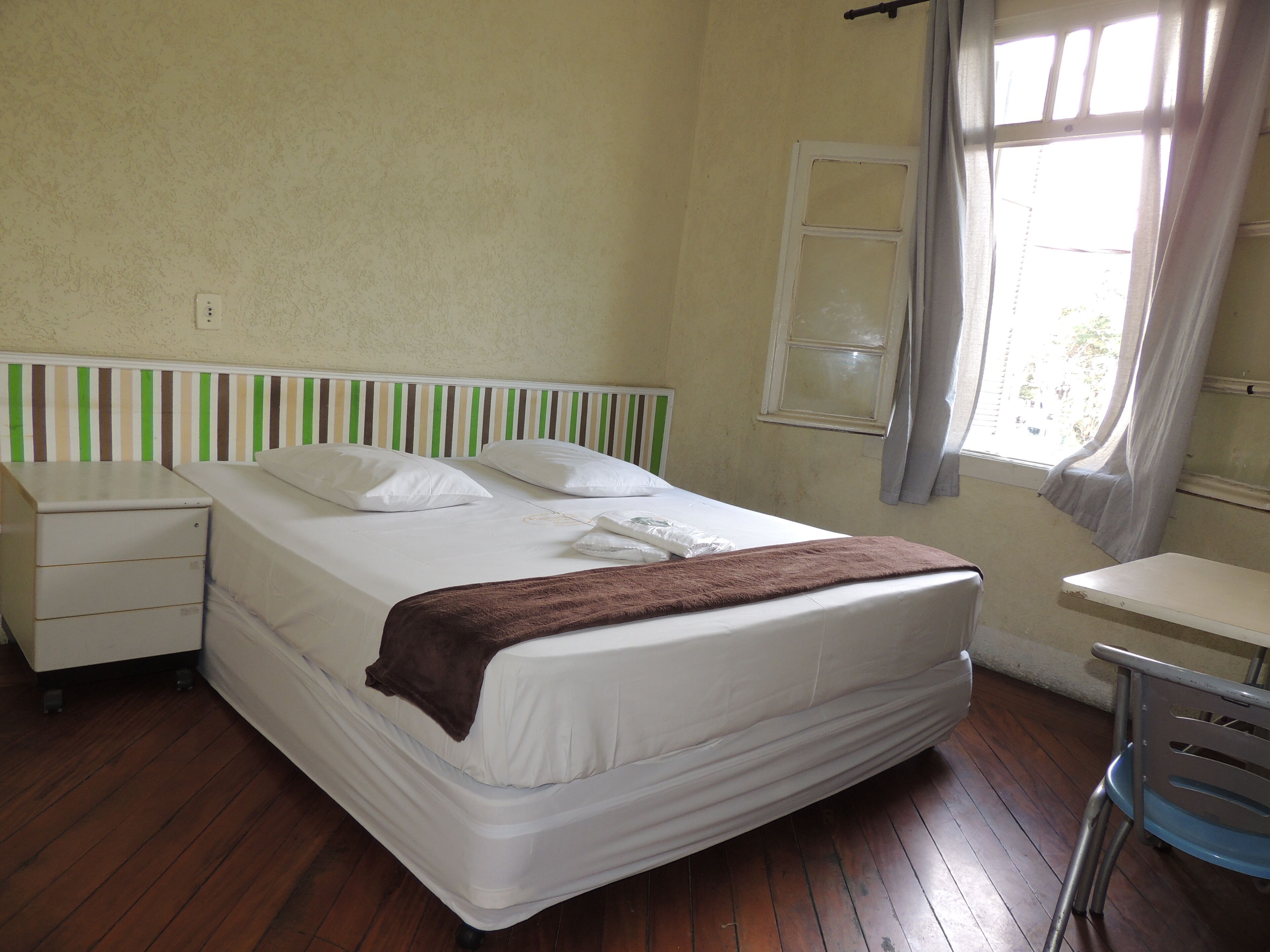 Habitación individual con piso de madera, mesita de noche, una silla, ventana con cortinas y cabecera a rayas.