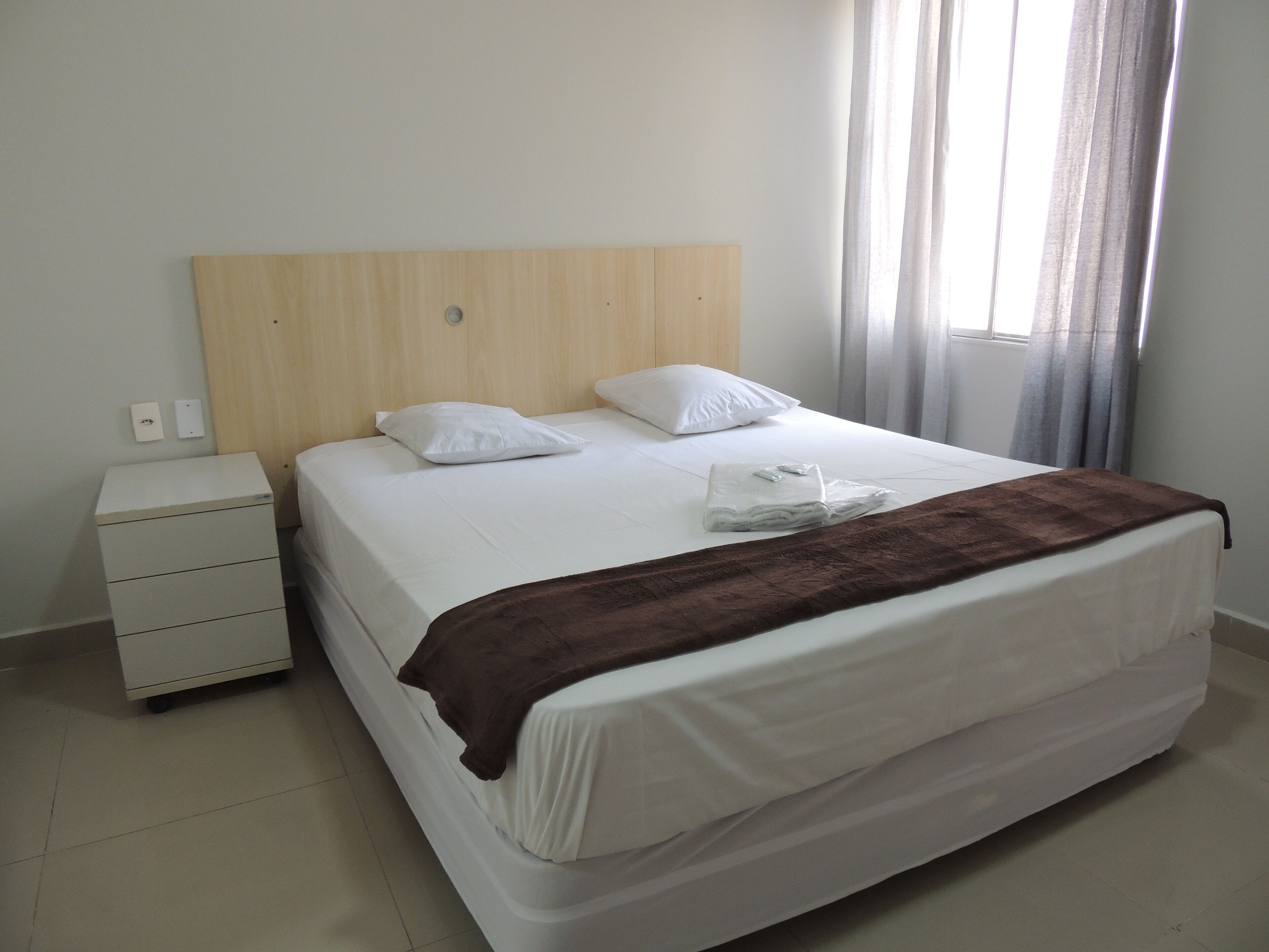 A neatly made bed with white linens and a brown throw blanket, a wooden headboard, and a small white bedside table with drawers.