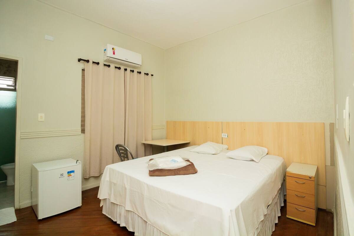 A hotel room with a bed, a desk, a chair, a small refrigerator, and a bedside table with drawers.