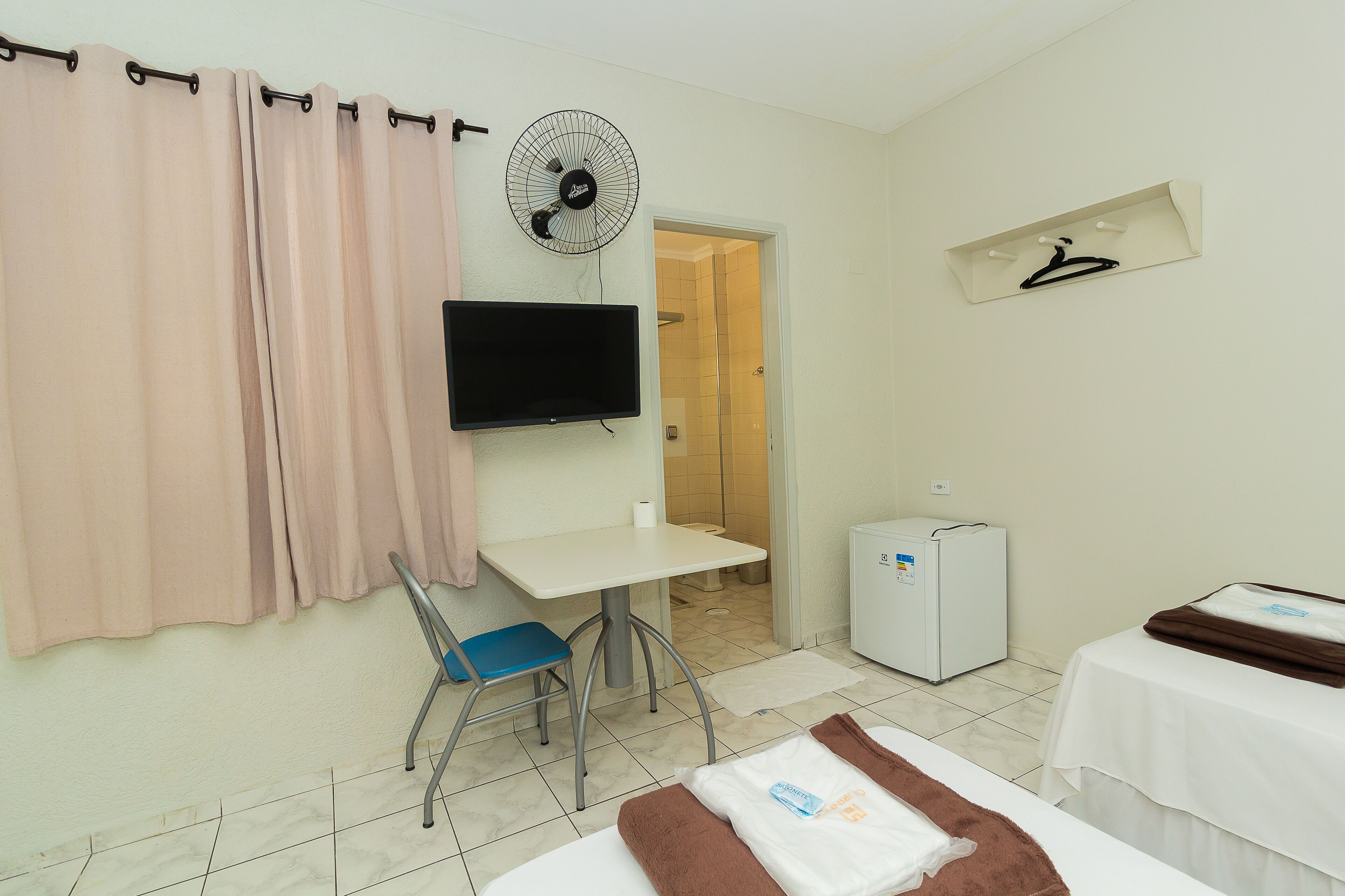 Una camera d'albergo con un letto, una scrivania, una sedia, una televisione, una ventola, un appendiabiti e un frigorifero.