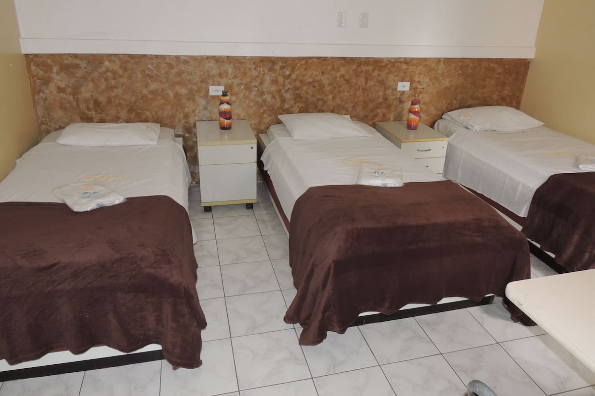 A hotel room with four single beds, each with a brown blanket and white bedding. There are two bedside tables with lamps and a small bottle on each. The room has a tiled floor and a textured wall above the beds.