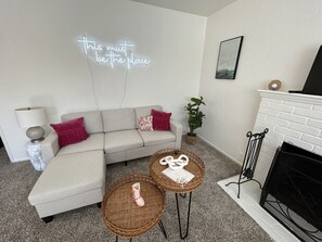 Una sala de estar moderna con un sofá blanco, un letrero de neón, una chimenea y una pequeña mesa con objetos decorativos.