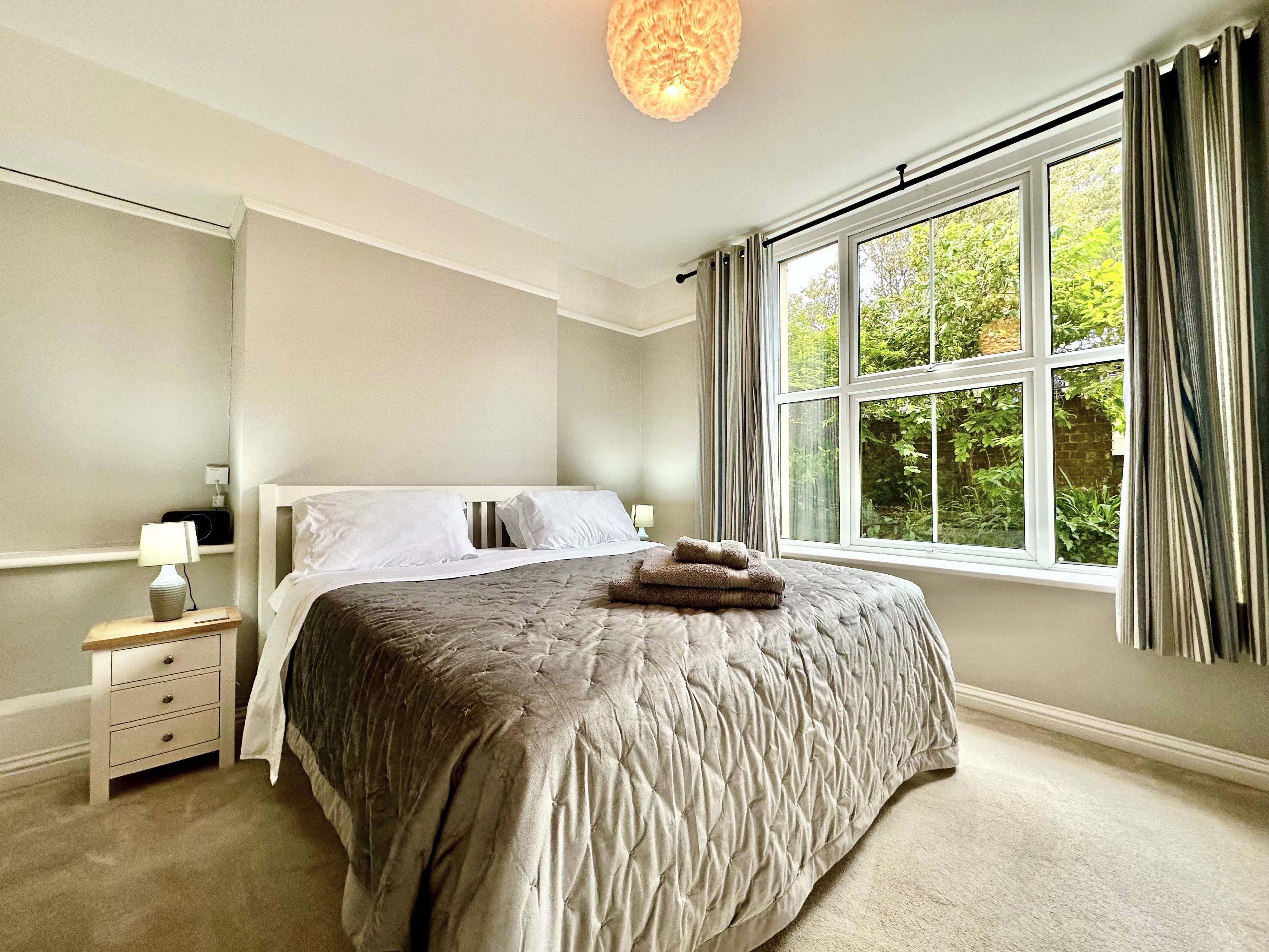 A bedroom with a large bed, a bedside table with a lamp, a window with curtains, and a view of greenery outside.