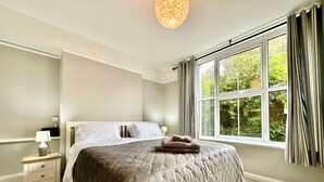 A bedroom with a large bed, a bedside table with a lamp, a window with curtains, and a view of greenery outside. - Smugglers View - Niton, sleeps 4 guests  in 2 bedrooms (Isle of Wight)