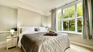A bedroom with a large bed, a bedside table with a lamp, a window with curtains, and a view of greenery outside.