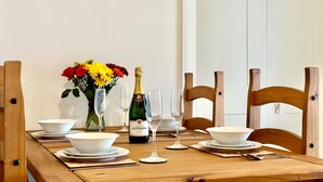 A dining table set with white bowls, plates, and cutlery, a bottle of champagne, and a vase of flowers. - Smugglers View - Niton, sleeps 4 guests  in 2 bedrooms (Isle of Wight)