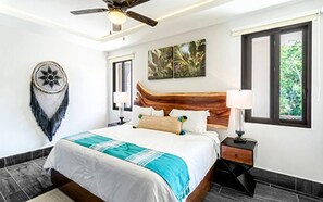 A modern hotel room with a wooden headboard, a large bed with a blue and white bedspread, a dreamcatcher on the wall, and a window with a view of greenery. - Bright Full Furnished 2 Bedrooms Waiting For You (Tulum)