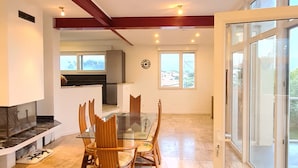 Un amplio salón con una mesa de comedor de vidrio, sillas de madera, una cocina al fondo y grandes ventanales que dejan entrar luz natural.