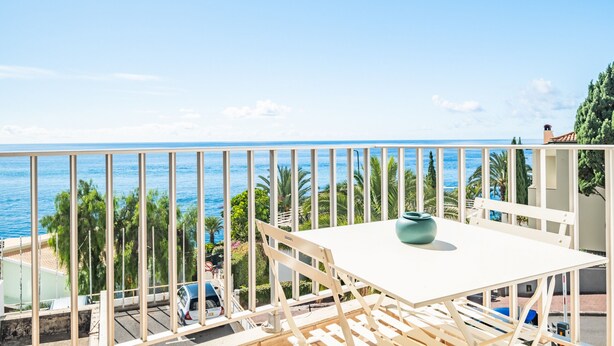 A balcony with a white table and chairs, overlooking a coastal view with palm trees and a clear sky.