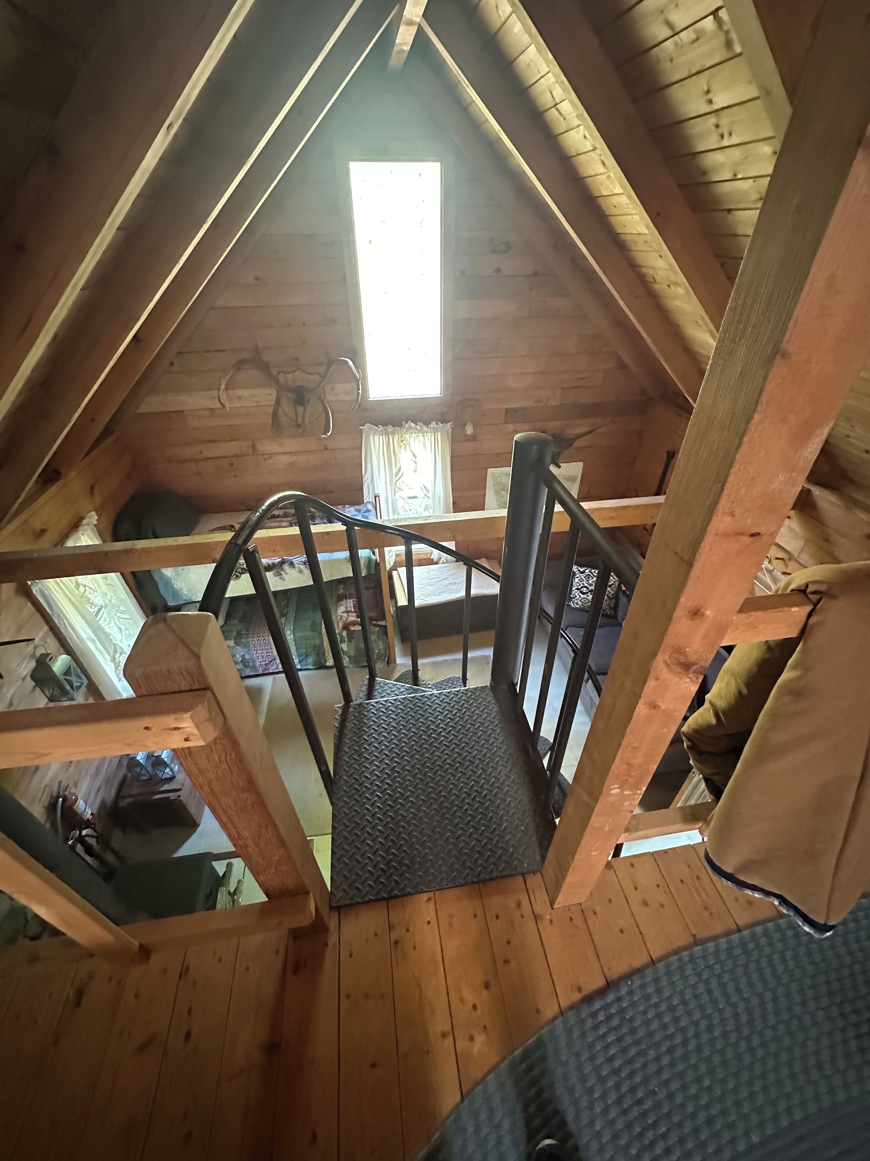A wooden loft bedroom with a sloped ceiling, a window with curtains, a metal ladder, and a bed with a patterned bedspread.