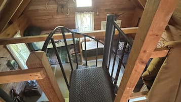 A wooden loft bedroom with a sloped ceiling, a window with curtains, a metal ladder, and a bed with a patterned bedspread.