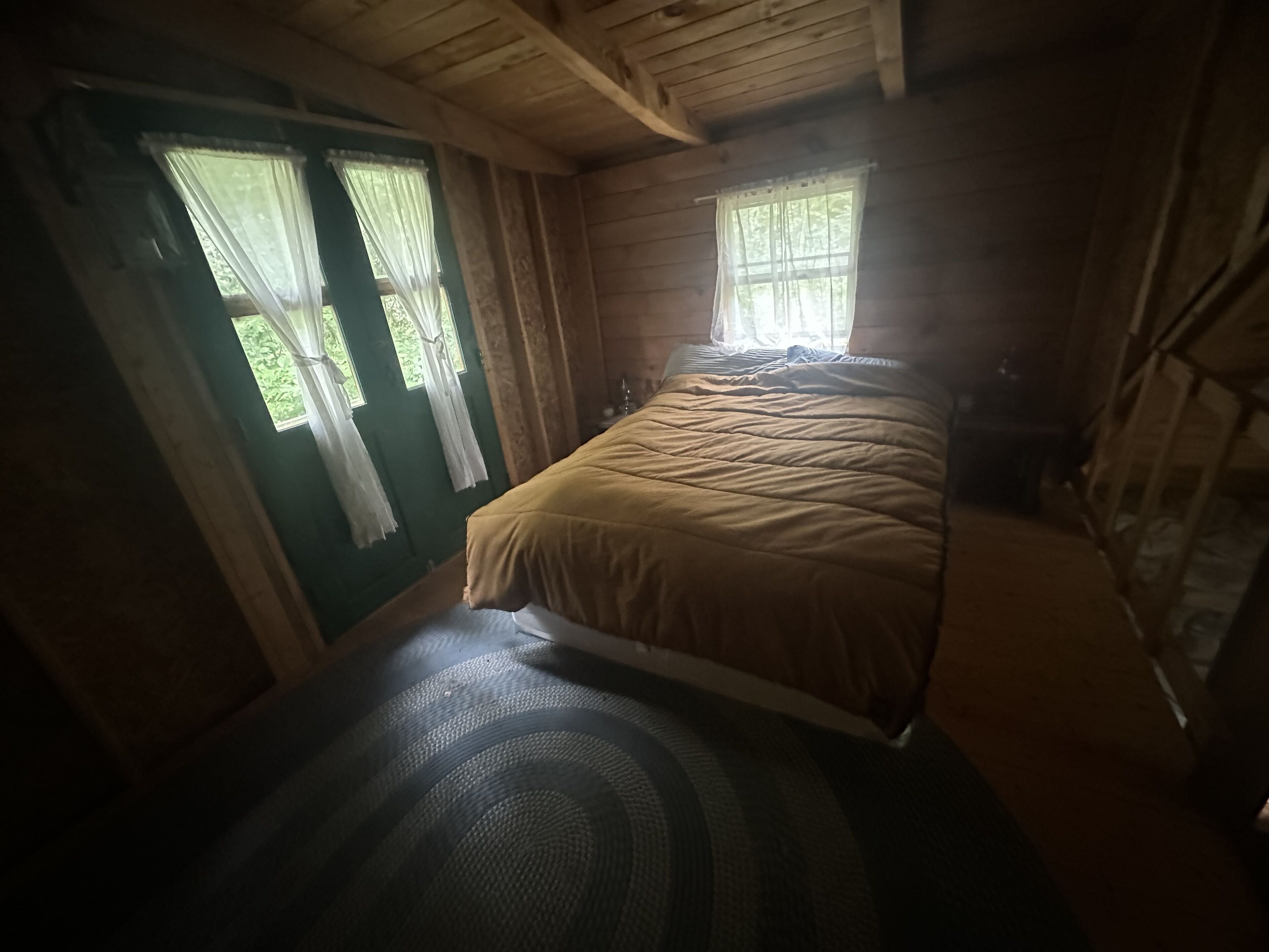 Un dormitorio de cabaña de madera con una cama, dos ventanas con cortinas y una puerta.