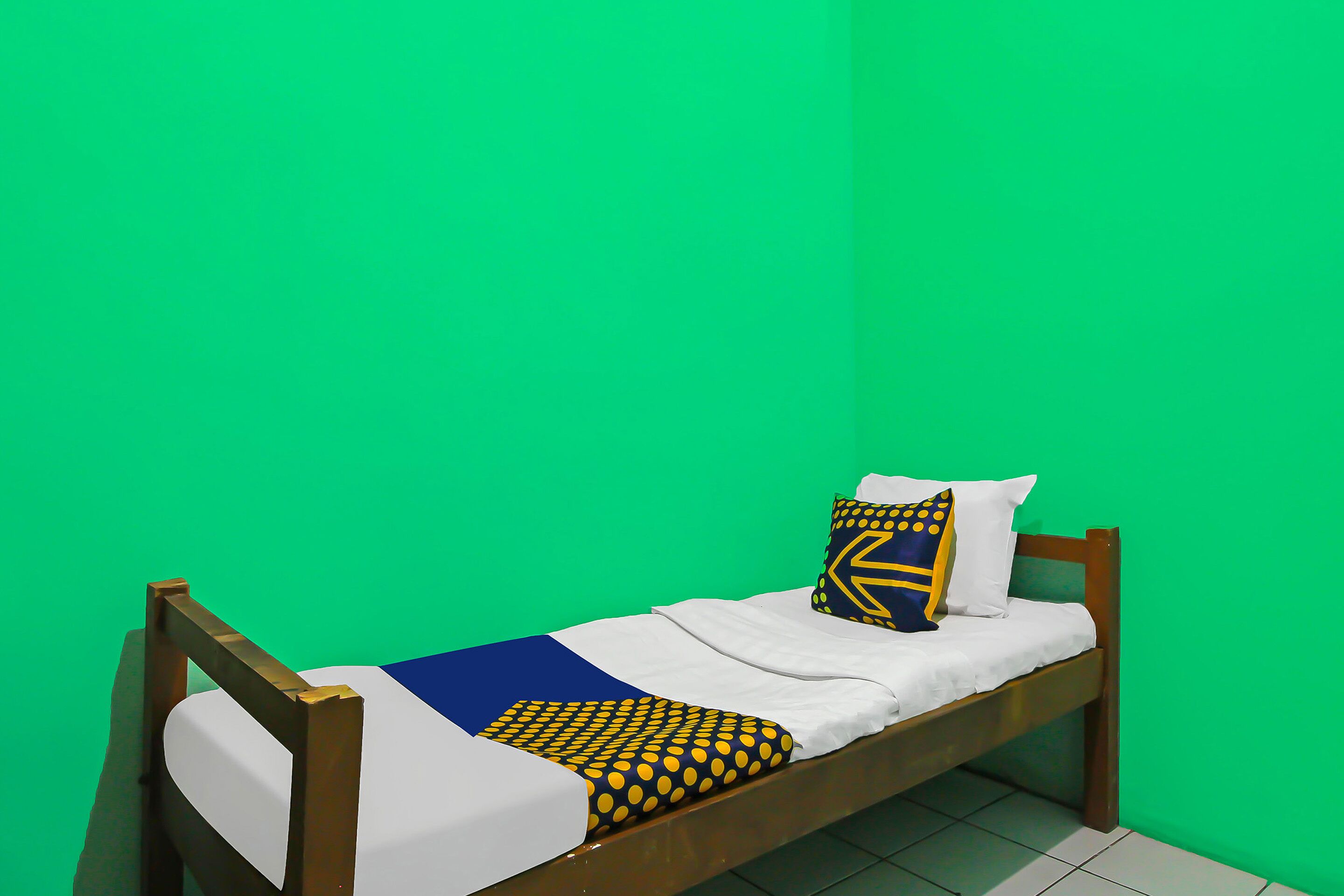 A single bed with a wooden frame, white bedding, and patterned pillows against a green wall.