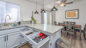 Eine moderne Küche mit weißen Schränken, Granit Arbeitsplatten und einer integrierten Kochinsel mit Schublade. Ein Deckenventilator, Pendelleuchten und ein Essbereich mit Holztischen und einer Tischplatte sind vorhanden.