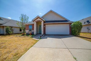 A two-story house with a front garage, a brick entrance, and a small front yard with a tree and shrubs. - Fenced Yard, Workspace, Garage: Ft Worth Home Base (Fort Worth)
