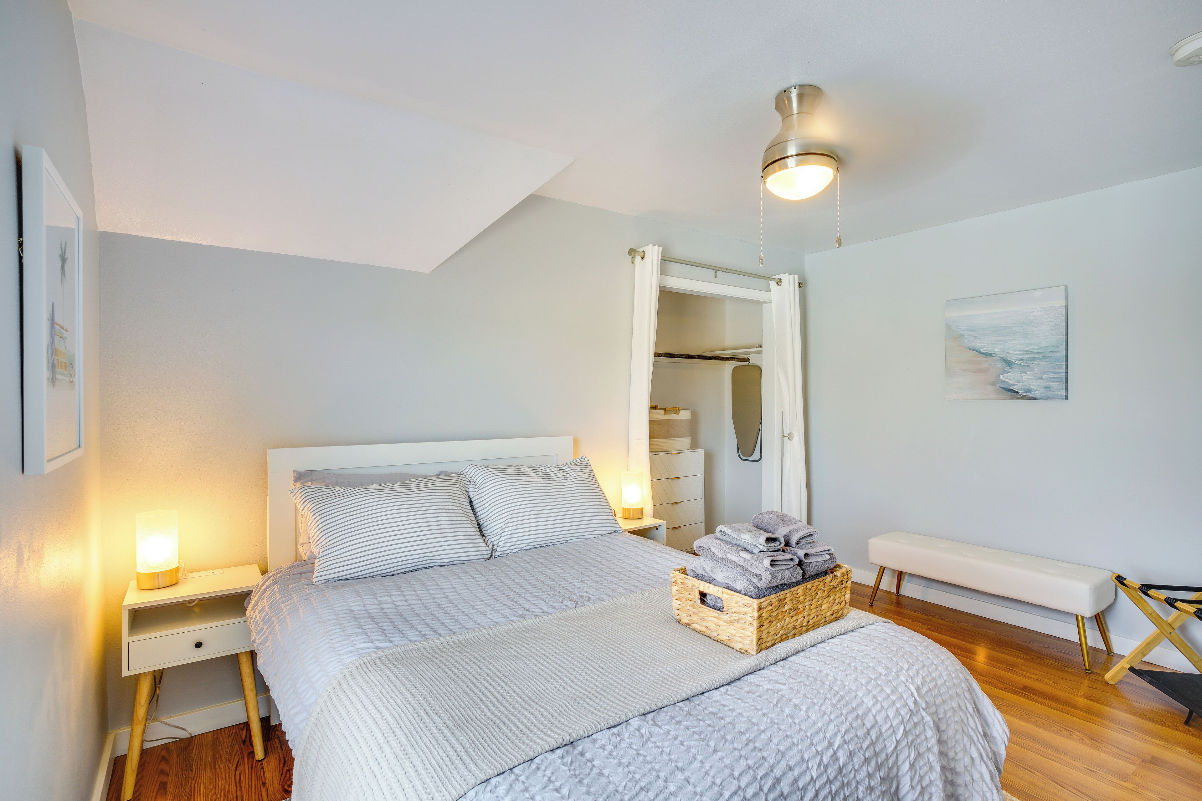 A bedroom with a bed, bedside lamps, a bench, a basket of towels, and a closet.