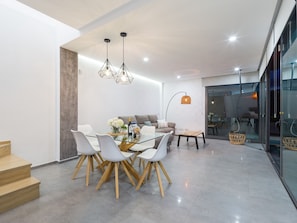 A modern dining area with a glass table, white chairs, and a sofa. There are two pendant lights above the table.