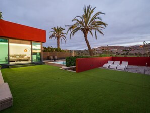 A modern house with a glass facade, a swimming pool, and a red wall with lounge chairs.