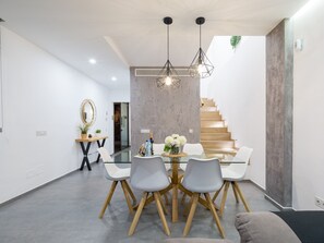 A modern dining area with a glass table, grey chairs, wooden legs, and hanging geometric light fixtures.