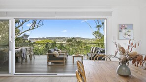 Ein geräumiger Balkon mit Holzterrasse, ausgestattet mit einer Couch, Stühlen und einem Esstisch. Eine große Schiebeglastür bietet Blick auf eine üppige Landschaft und sanfte Hügel in der Ferne.