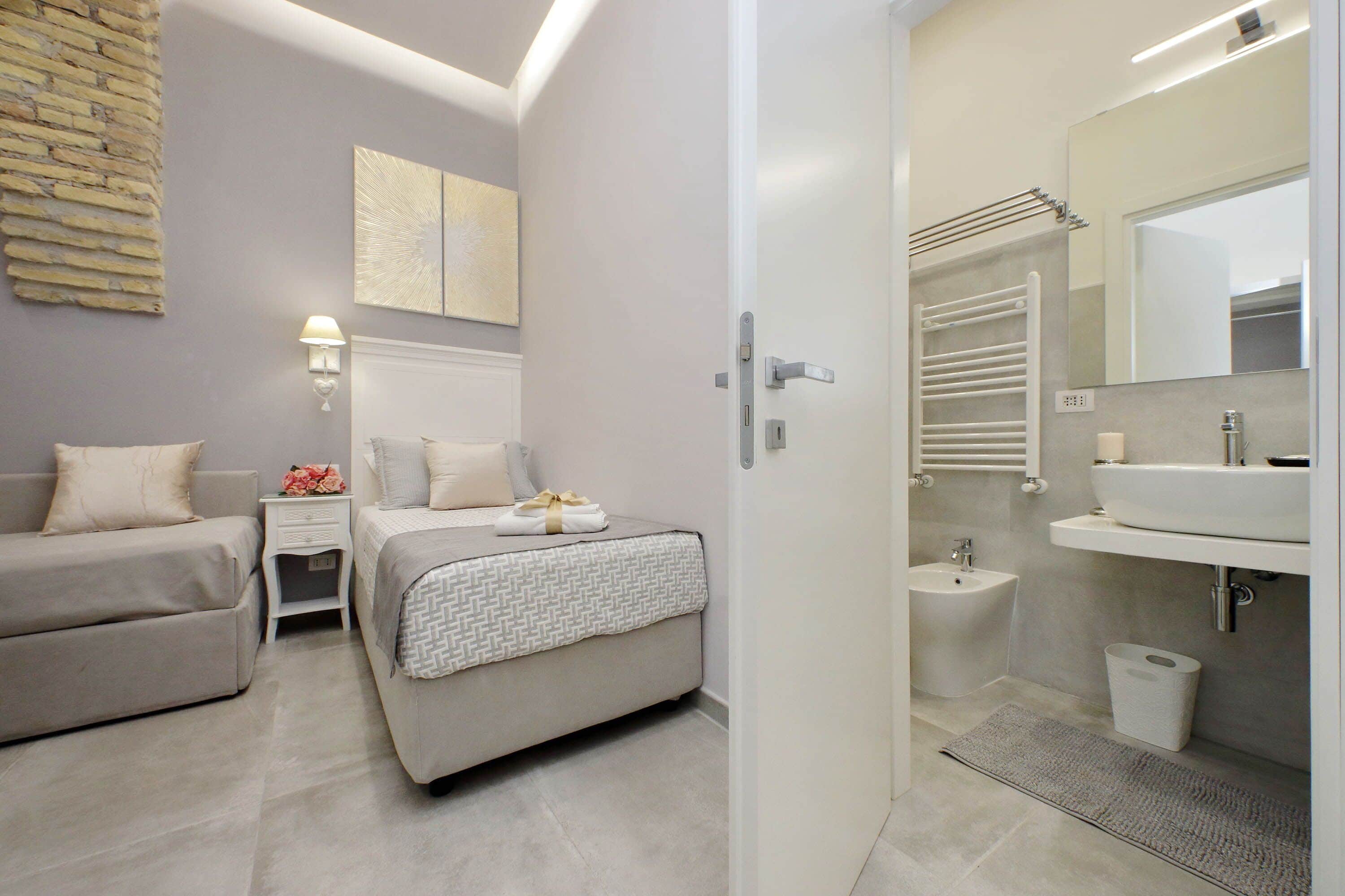 A modern bedroom with a bed, bedside tables, a nightstand, and a bathroom with a sink, mirror, and towel rack.