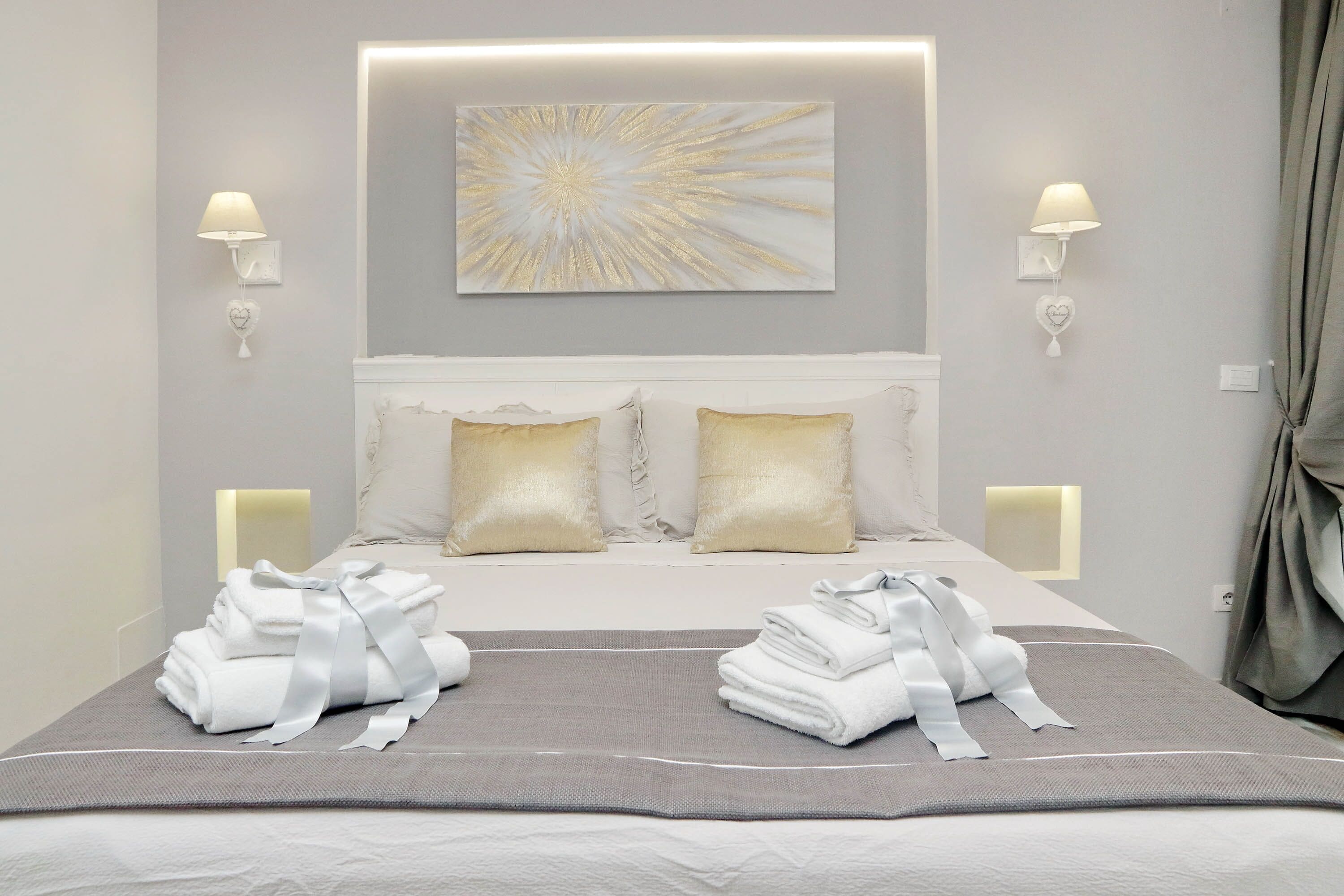 Una cama bien hecha con sábanas blancas y una manta gris, dos toallas con un lazo, y una obra de arte abstracto colgada en la pared.