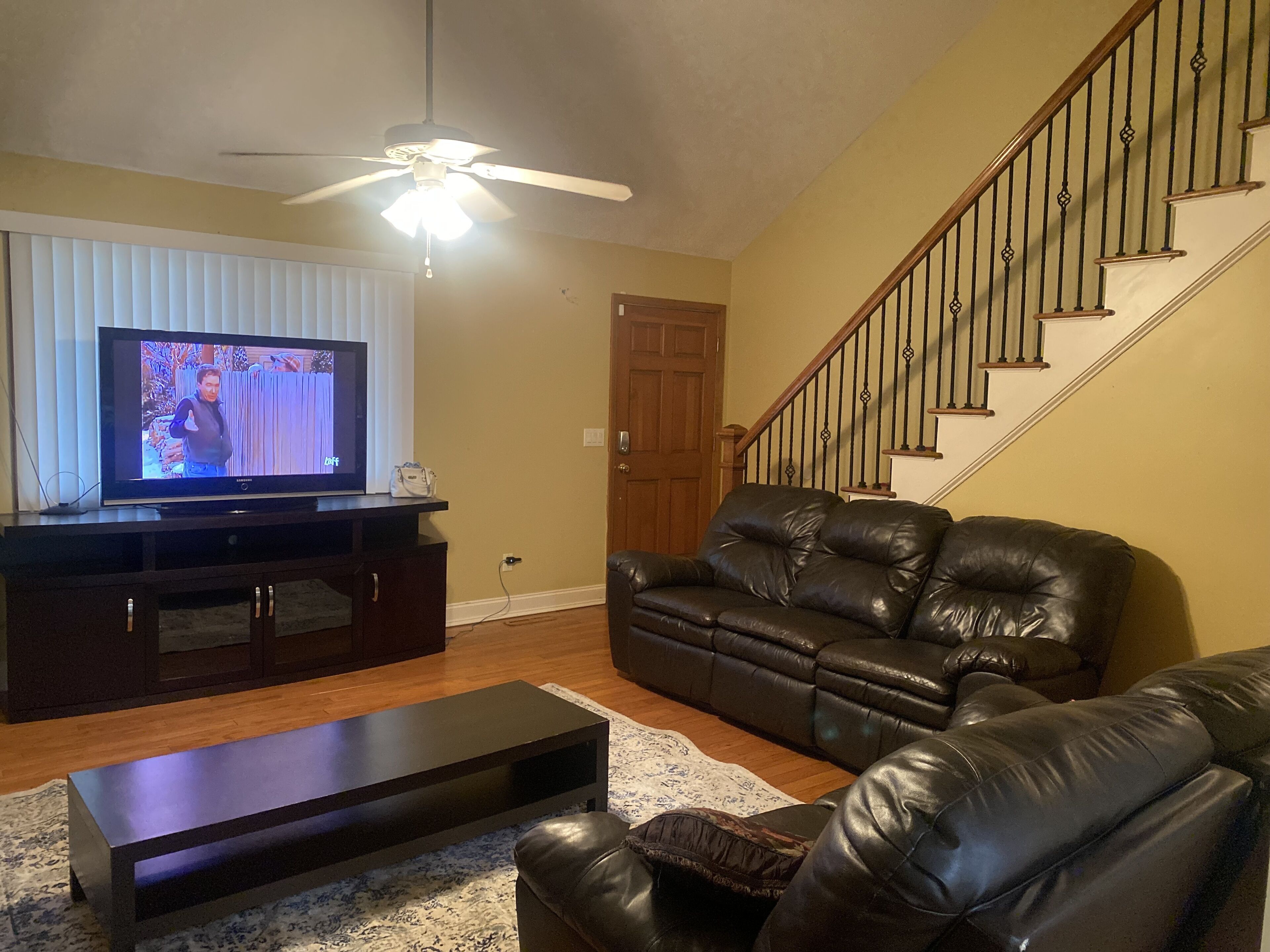 Ein Wohnzimmer mit Fernseher, Ledersofa, Couchtisch, Deckenventilator und einer Treppe.
