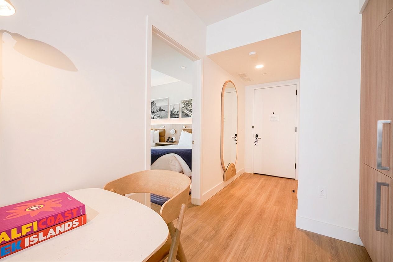 A hotel room with a wooden floor, a round table with a chair, a bed with a blue bedspread, a white door, and a mirror.