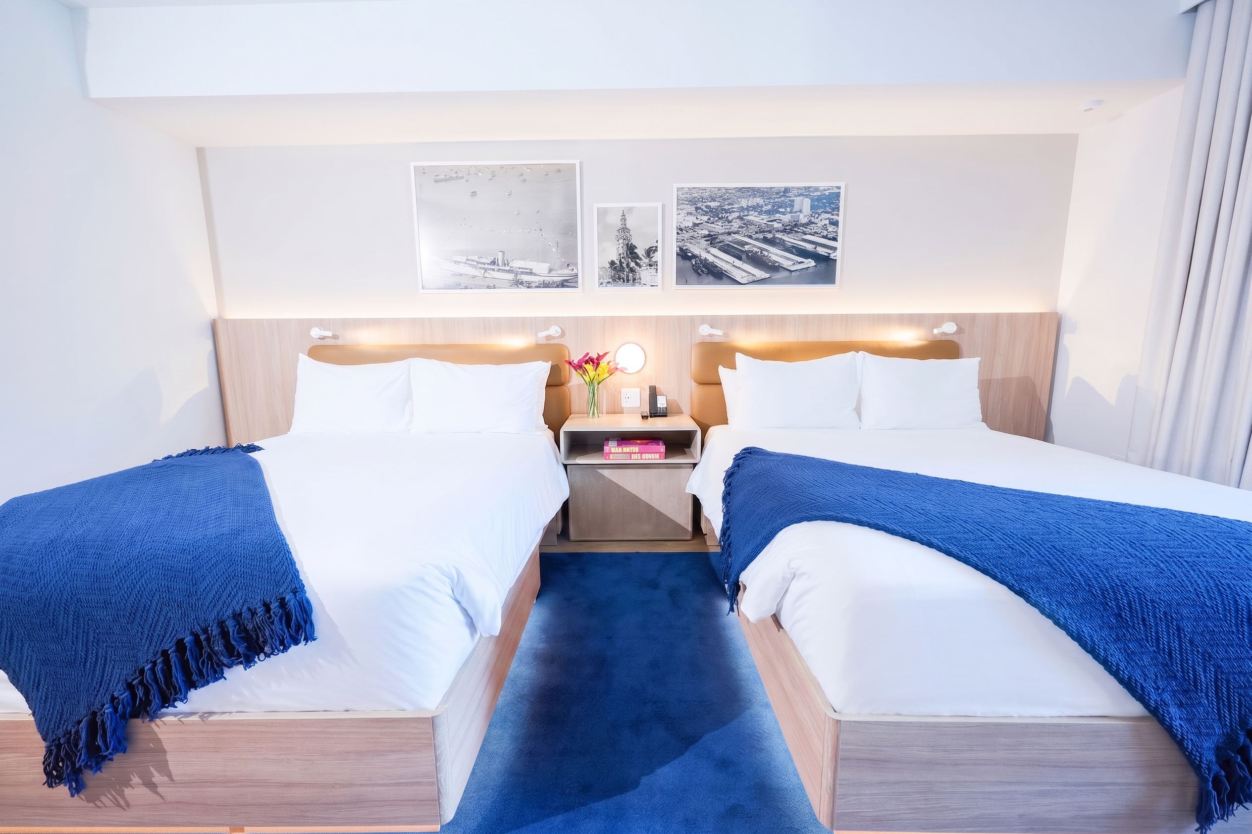 Habitación de hotel con dos camas, una mesita de noche con lámpara y dos cuadros en la pared.