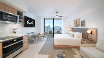 A modern hotel room with a kitchenette, a bed with a city view, a seating area, and a flat-screen TV.