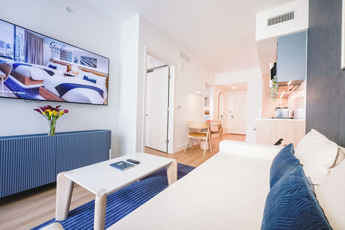 A modern hotel room with a bed, a TV, a dining area, and a kitchenette.