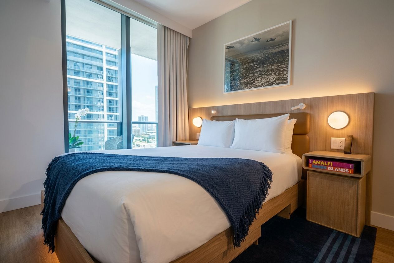 A hotel room with a large bed, a nightstand with a book, a window with a view of buildings, and a painting on the wall.