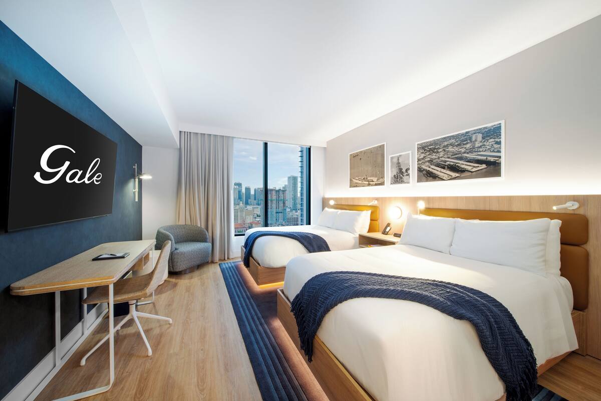 A hotel room with two beds, a desk, and a large window offering a city view.