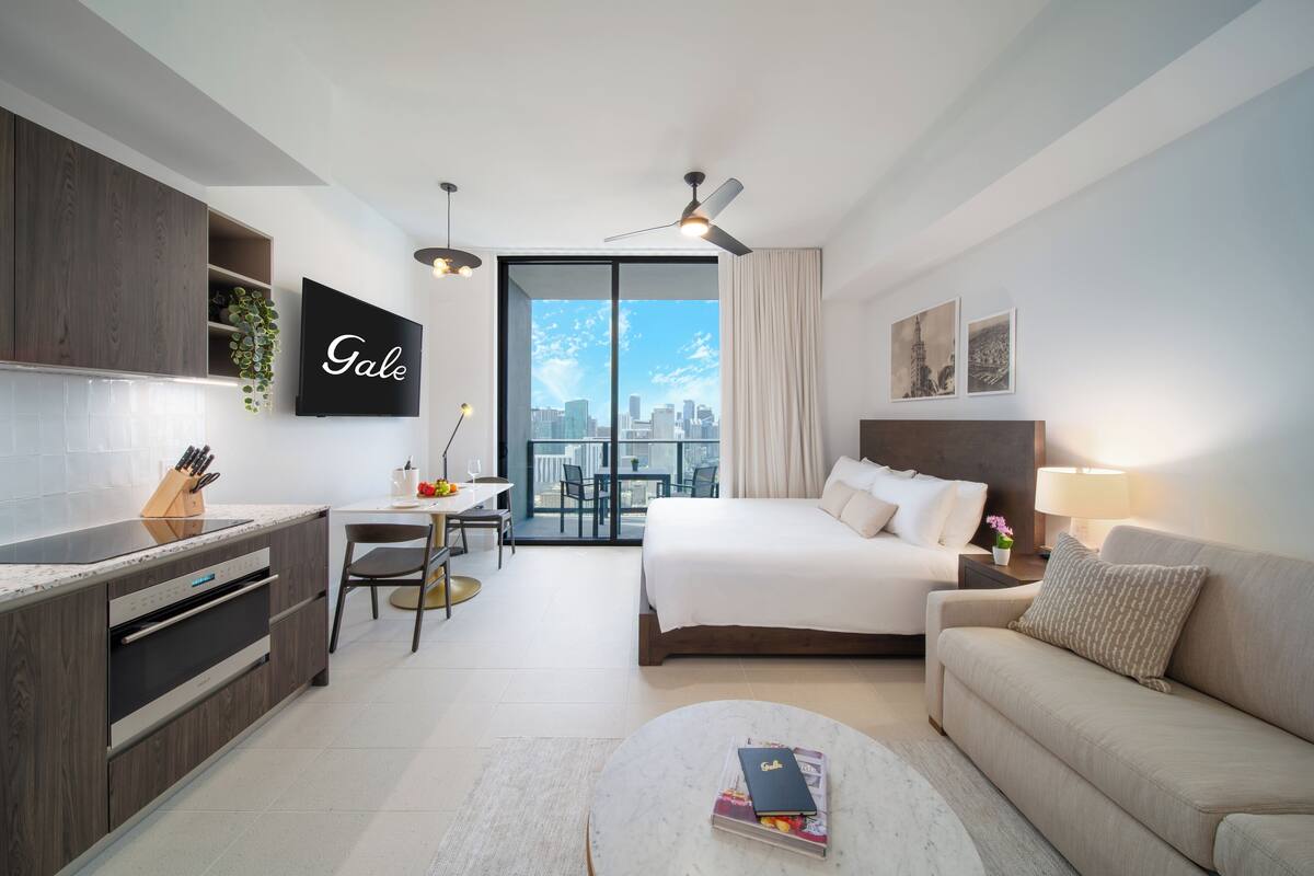 A modern hotel room with a balcony, a bed, a sofa, a dining area, and a kitchen.