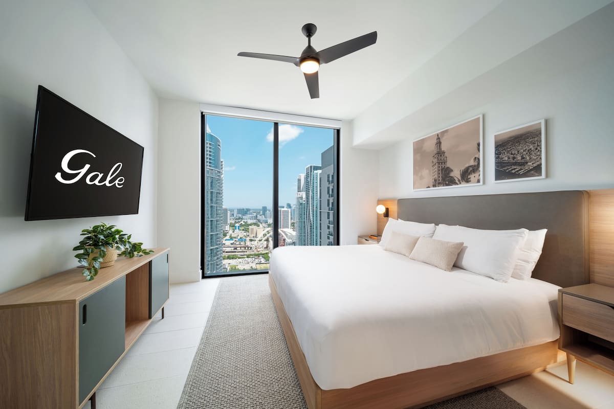 A modern hotel room with a large bed, a ceiling fan, a TV mounted on the wall, and a city view through the sliding doors.