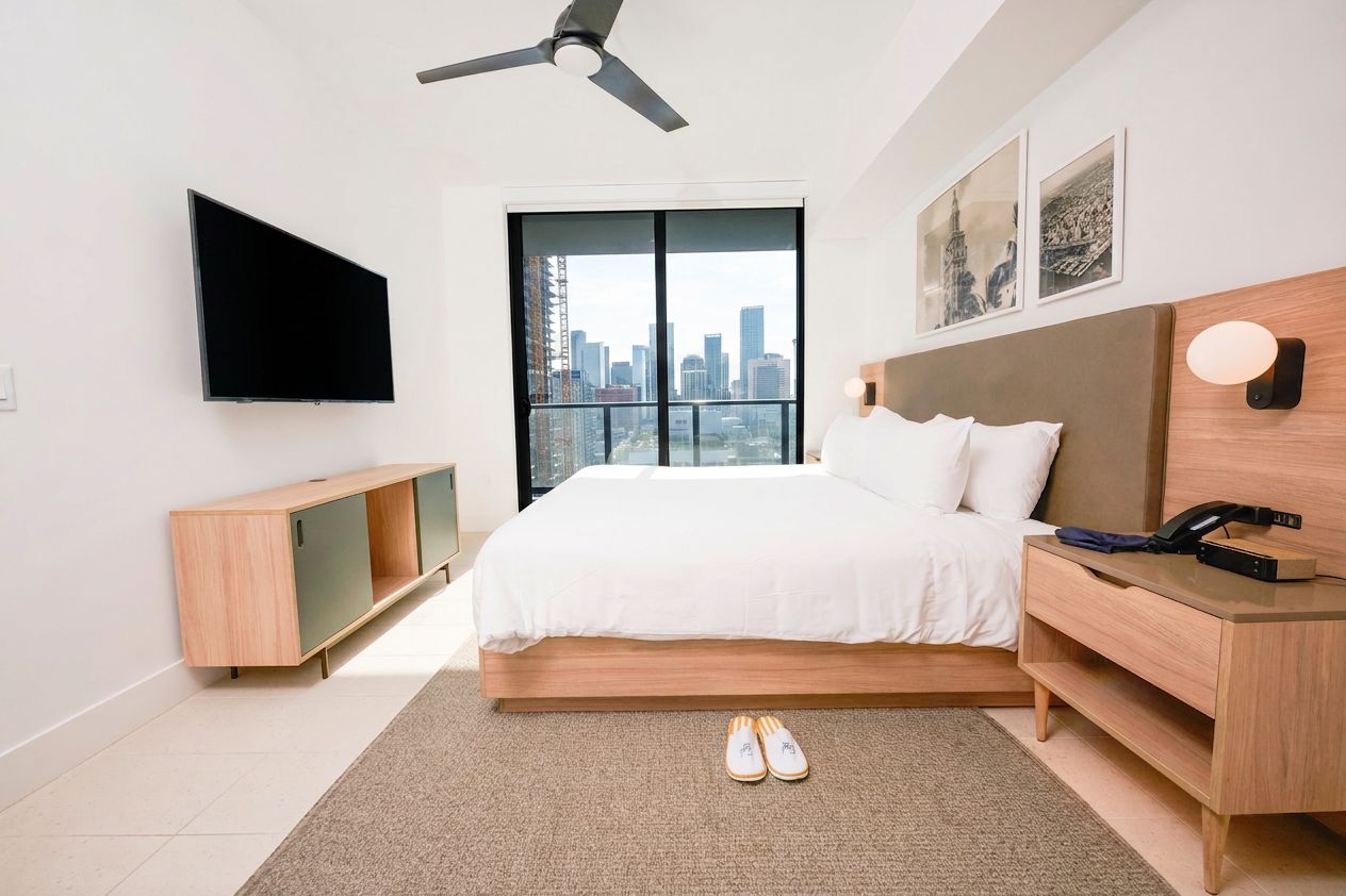 Un dormitorio moderno con cama, televisión, mesita de noche, escritorio y vista a la ciudad.