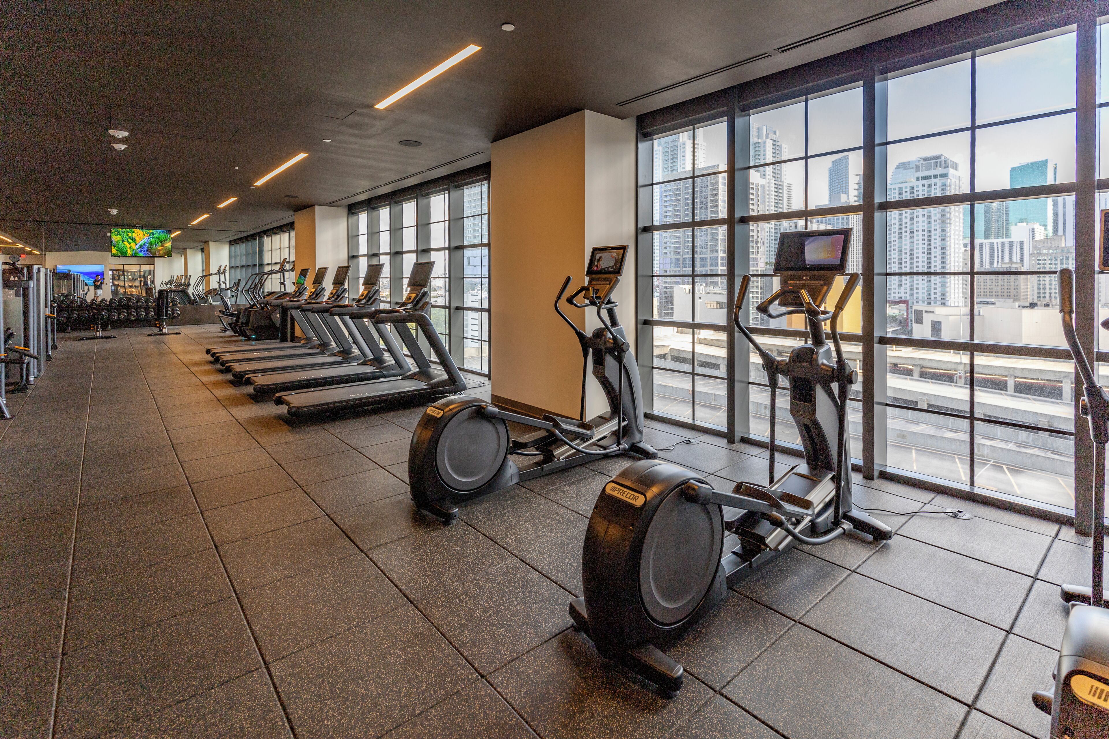 A gym with treadmills and elliptical machines, large windows, and a city view.