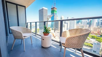 En balkon med to fletstole, en potteplante og udsigt over et bybillede med høje bygninger.