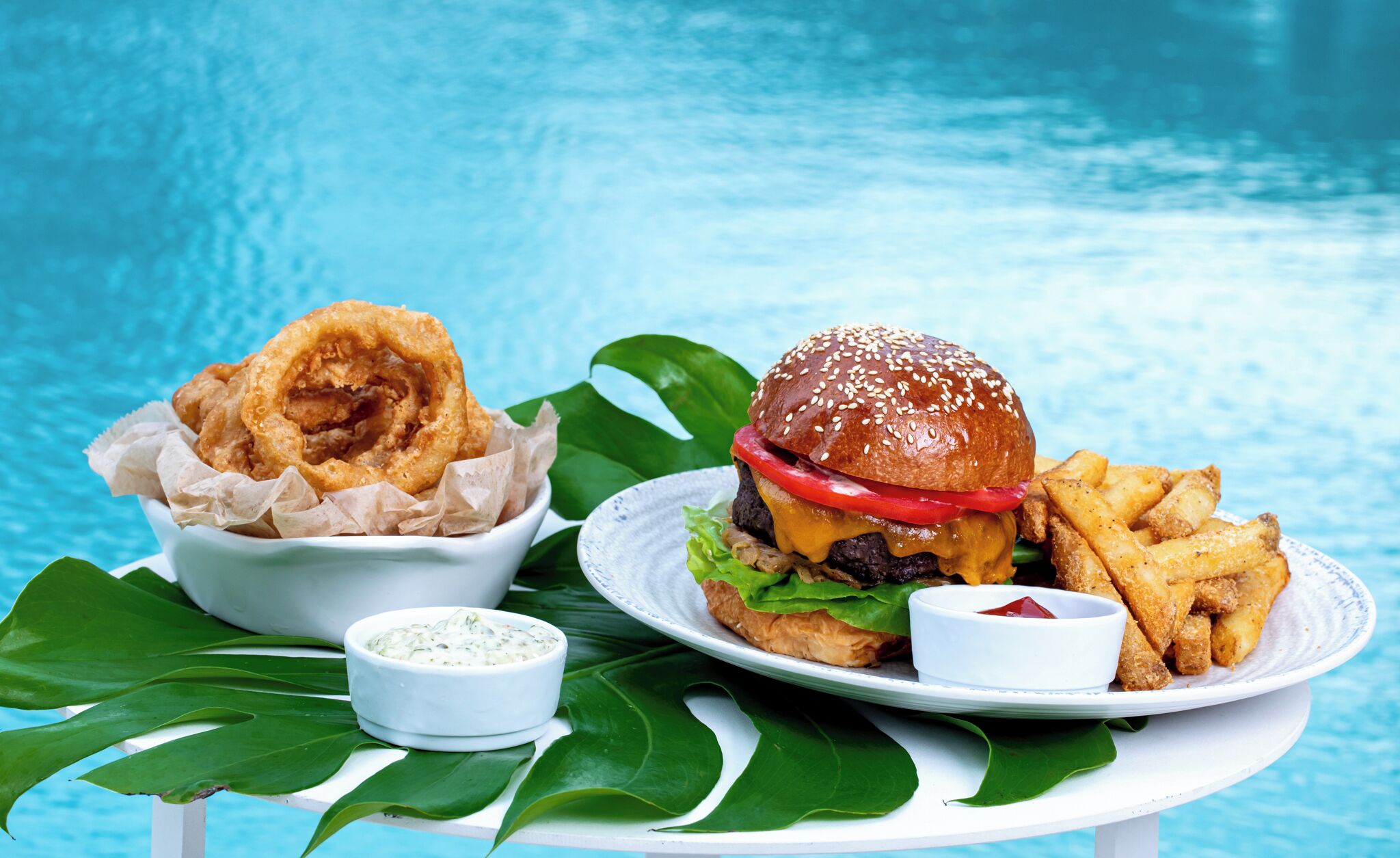 Ein Burger mit Pommes und Zwiebelringen auf einem weißen Teller, begleitet von einer kleinen Sauce, alles auf einem Tisch mit blauem Hintergrund platziert.