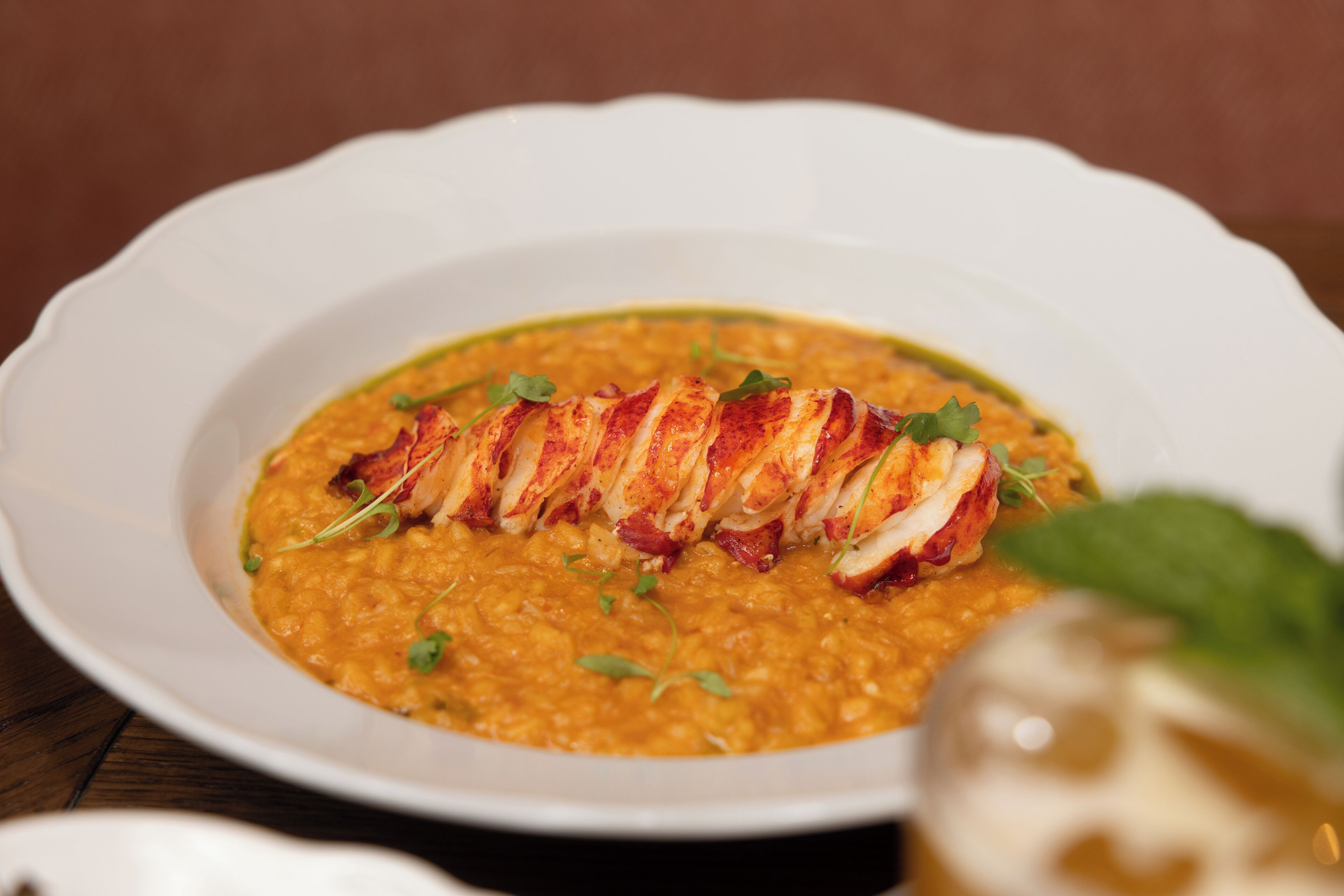 Un tazón blanco con una cola de langosta sobre una sopa cremosa anaranjada, adornado con hierbas verdes.