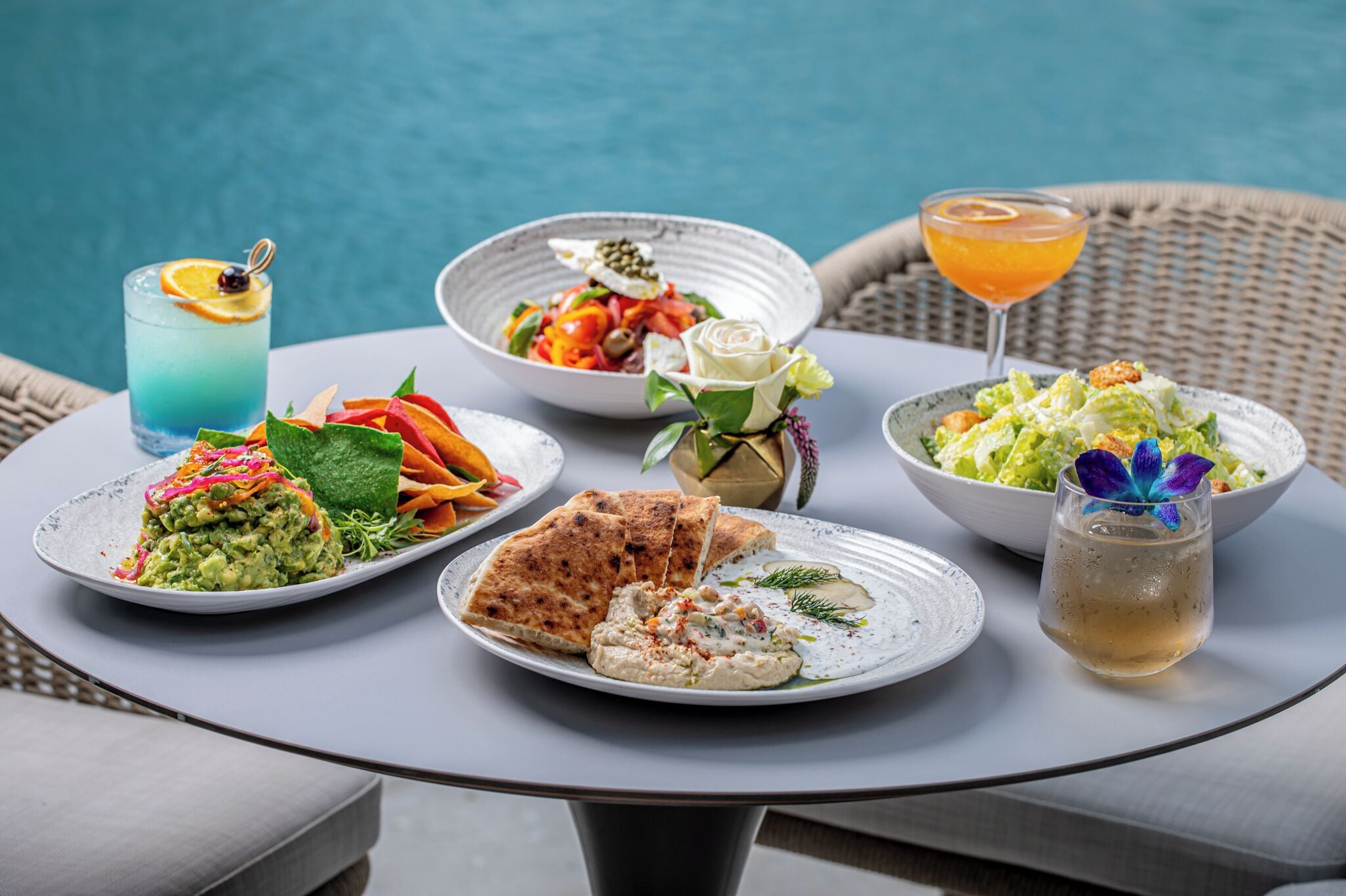 A table with various dishes including a blue drink, a salad, and a bowl of food, set against a poolside view.
