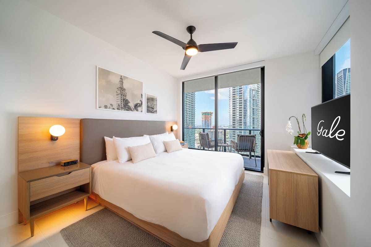 A modern hotel room with a large bed, a ceiling fan, a desk, and a balcony with a city view.