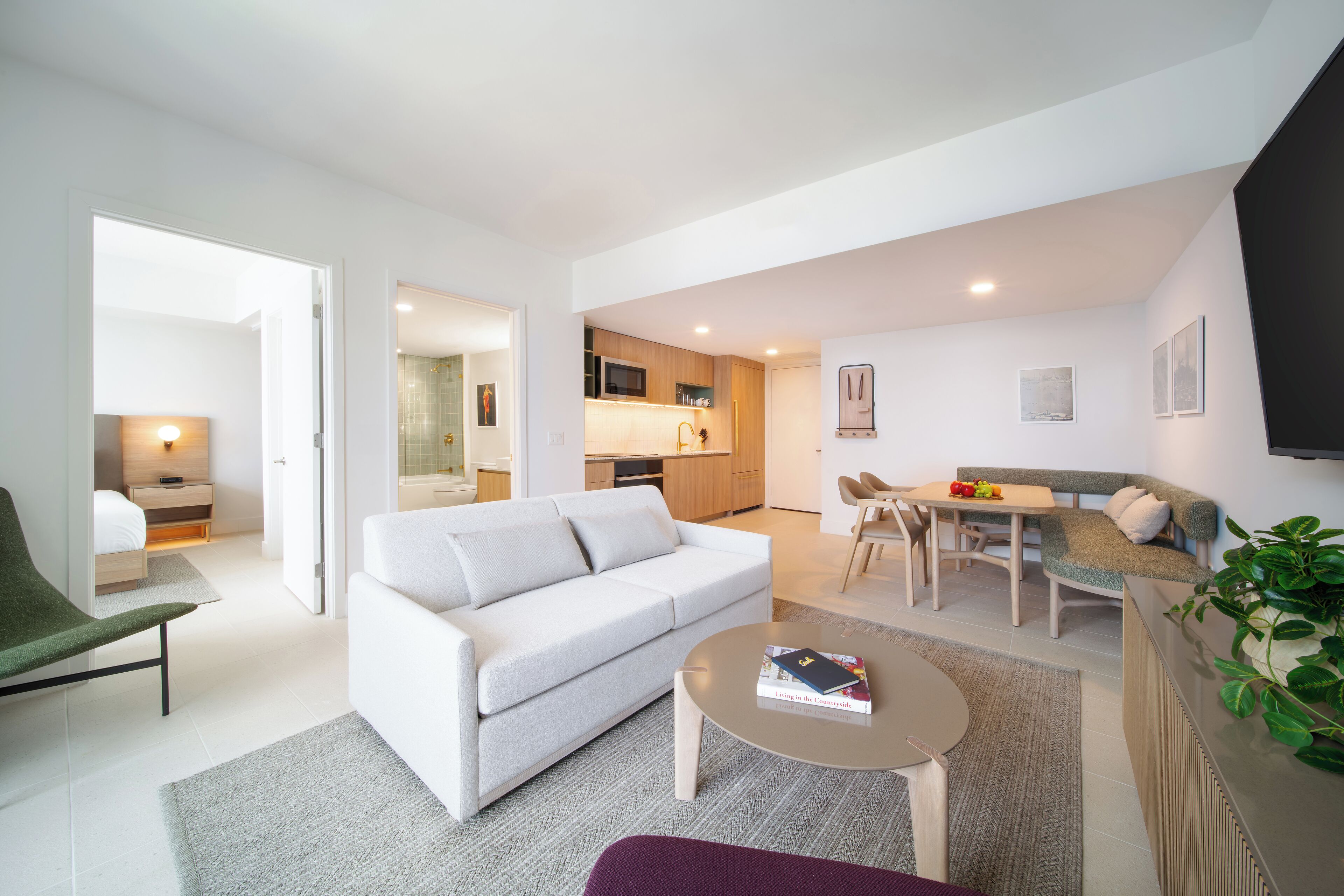 Un salon moderne avec un canapé blanc, une table à manger avec des chaises, un coin cuisine et un téléviseur à écran plat fixé au mur.