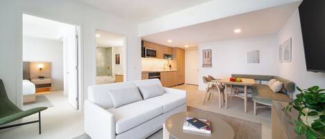 A modern living room with a white sofa, a dining table with chairs, a kitchen area, and a flat-screen TV mounted on the wall.