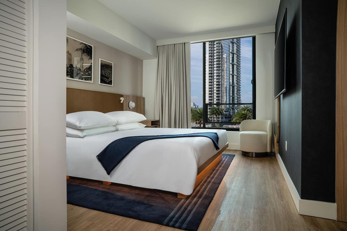 A modern hotel room with a large bed, a chair, a TV, and a view of tall buildings.