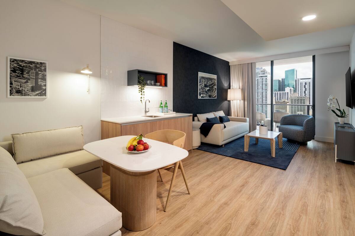 A modern living room with a sofa, a dining table with fruit, a kitchen area, a balcony with a view of the city, and a TV mounted on the wall.
