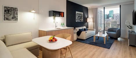 Una sala de estar moderna con sofá, una mesa de comedor con fruta, una zona de cocina, un balcón con vistas a la ciudad y un televisor montado en la pared.