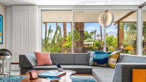 A modern living room with a grey sectional sofa, a coffee table, and a view of palm trees and mountains through large windows.